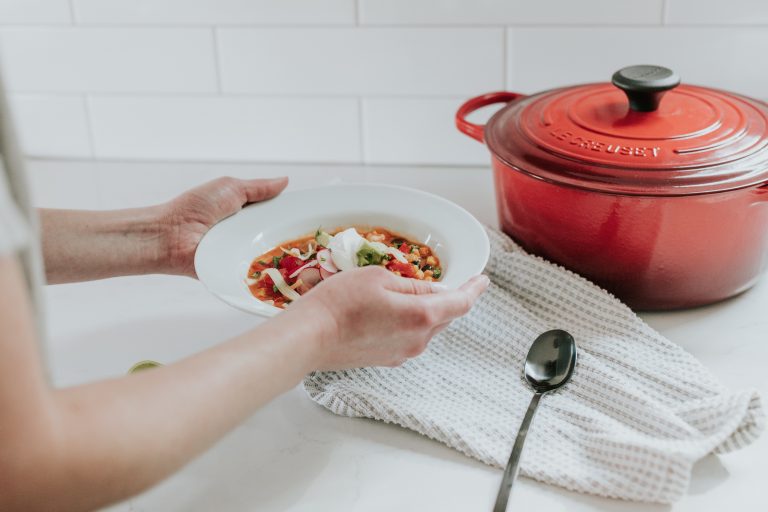 a cropped image of a person lifting a white bowl full of soup from a white counter with a red dutch oven next to it