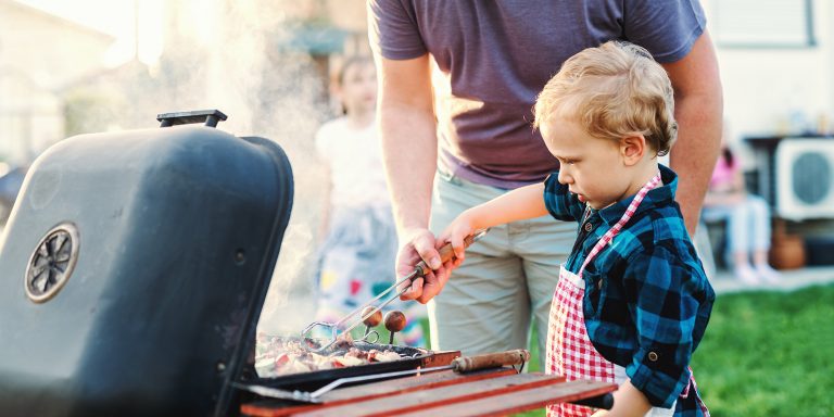a little boy helps his father flip hotdogs on the grill, wearing a checkered apron