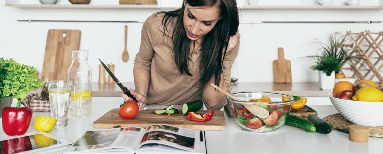 a woman leaning over a cutting board with a knife in hand, reading a recipe from acookbook