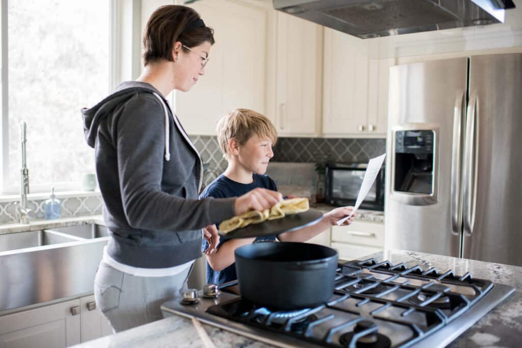 a woman holds open the lid of a pot, while son hold up recipe instructions on a piece of paper