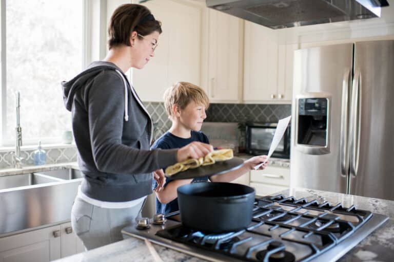 a woman holds open the lid of a pot, while son hold up recipe instructions on a piece of paper