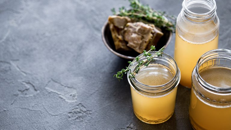Glass jar with yellow fresh bone broth on dark gray background. Healthy low-calories food is rich in vitamins, collagen and anti-inflammatory amino acid