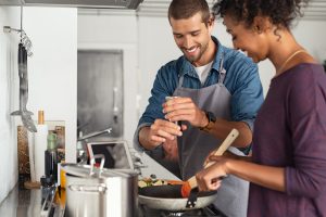 Man adding pepper to tomato sauce, while woman stirs with wooden spoon