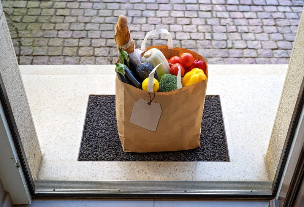 Contactless food delivery service concept. Paper bag with groceries delivered and left outside at entrance door. View from inside through open door. Online shopping. Zero waste package