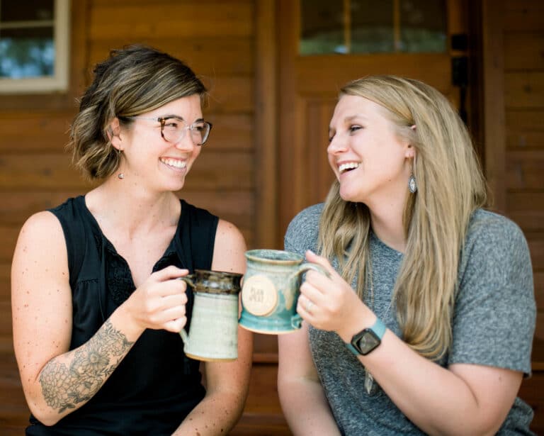 two young women smiling and clinking mugs together outside on a front porch