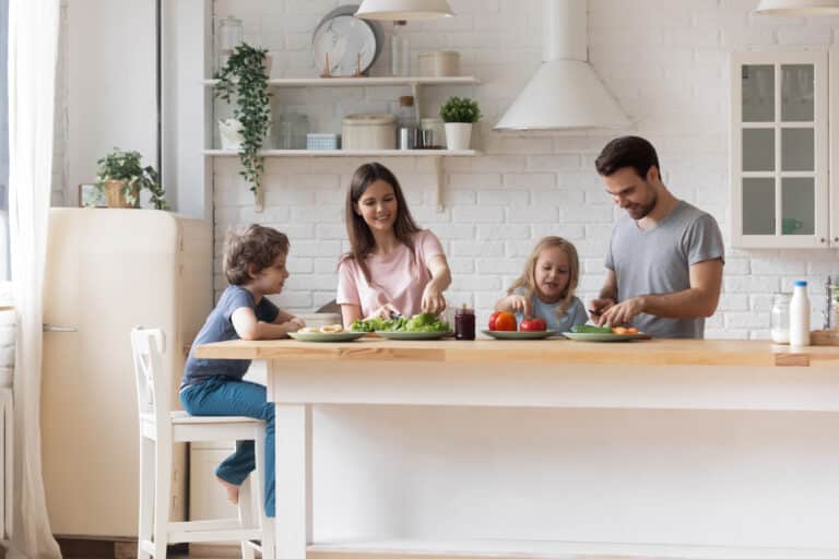 Happy parents with little children preparing salad for dinner together, standing at table at home, talking, smiling mother and father cutting vegetables in modern kitchen, family enjoying weekend