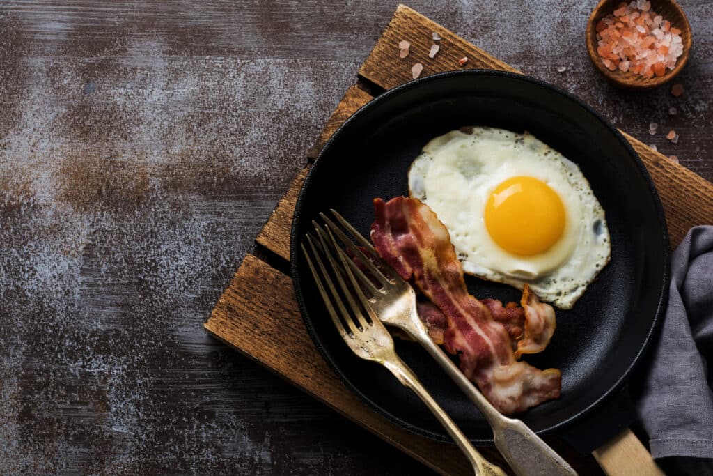 Traditional English breakfast with fried eggs and bacon in cast iron pan on dark concrete background. Top view.