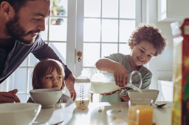Man watching his kid pour milk in his breakfast bowl. Father and kids sitting at the table preparing breakfast.
