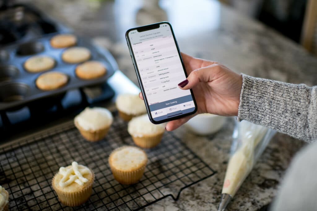 cropped image of a woman's hand holding a phone with a recipe visible, while frosting cupcakes