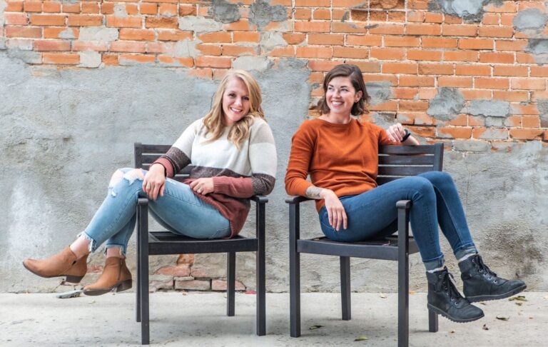 Riley and Roni sitting sideways on black metal chairs with a brick wall behind them