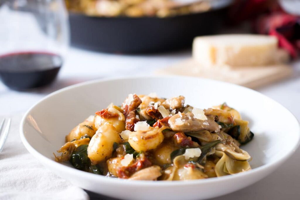 Angled photo of chicken gnocchi recipe in a white bowl with a wine glass and cheese in the background