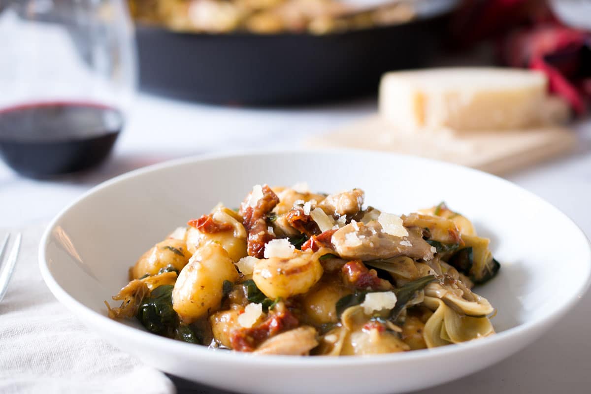 Angled photo of chicken gnocchi recipe in a white bowl with a wine glass and cheese in the background