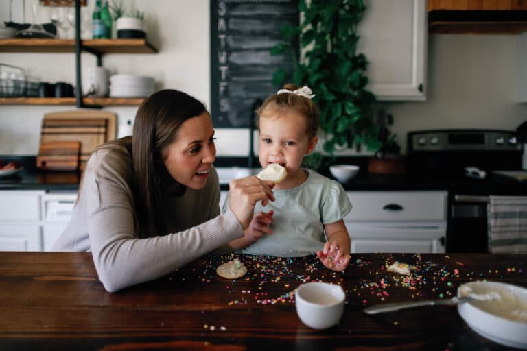 a mother helping her daughter eat a bite of a sugar cookie at a kitchen island