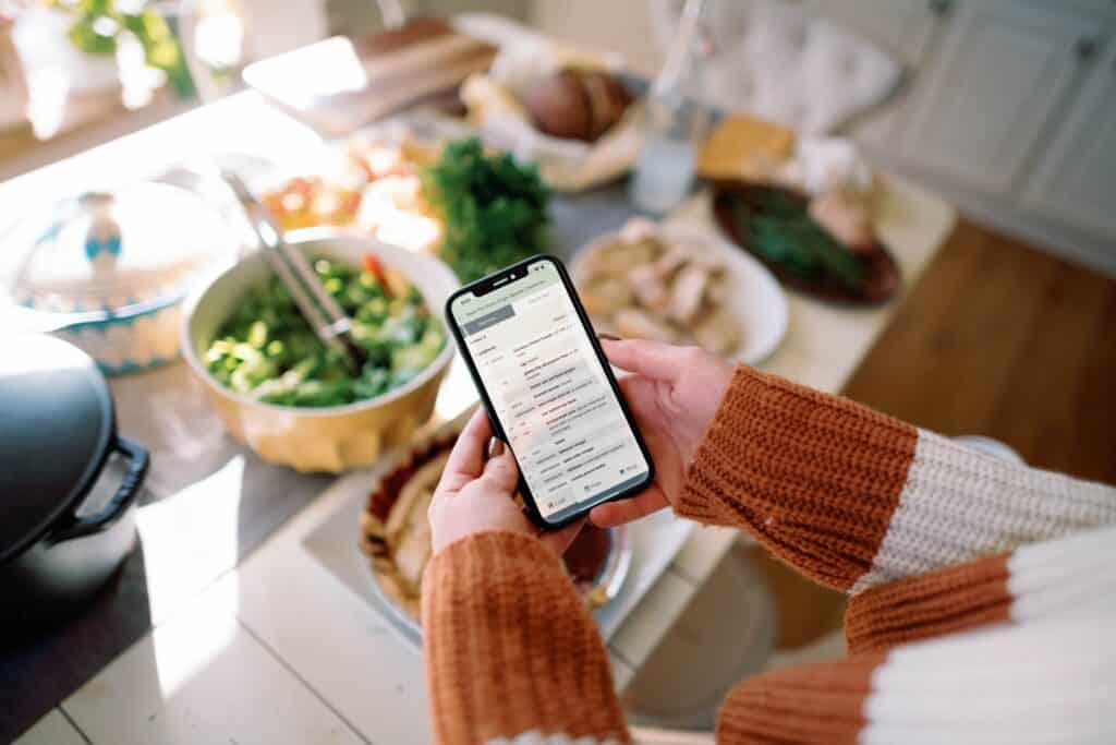 cropped image of a person viewing a recipe on their phone with a table full of food in the background.