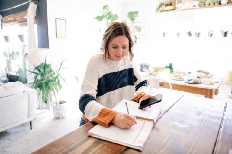 woman sitting at a dining table writing on a planner with a cell phone in her hand