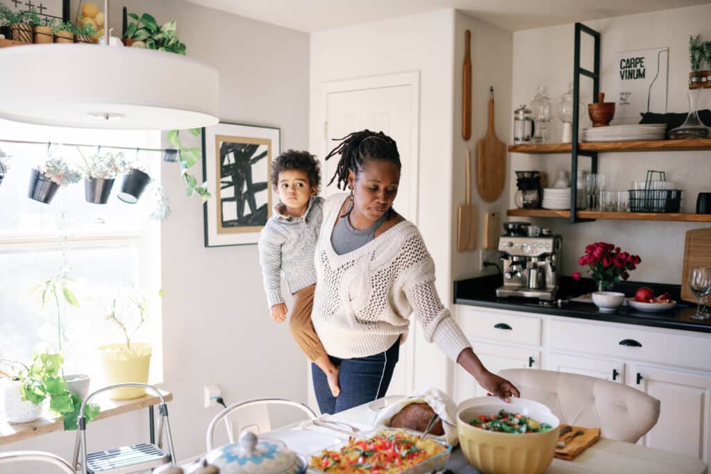 young black woman putting place settings on a dinner table while holding child.