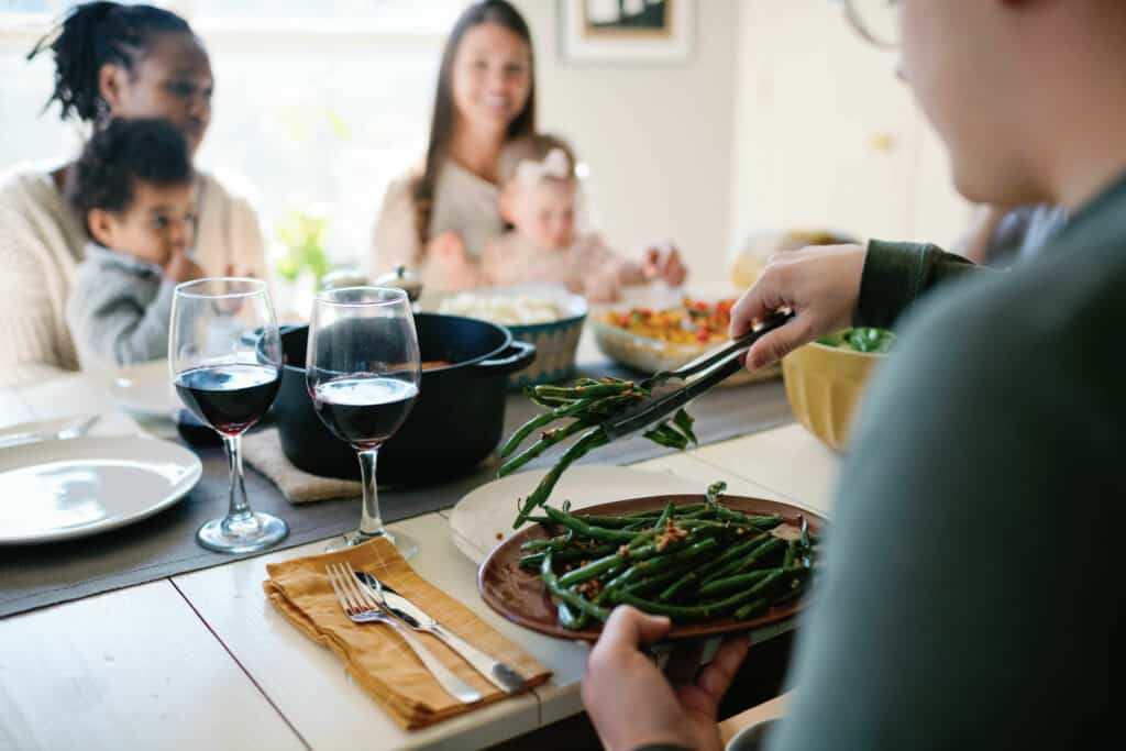 over the shoulder shot of a man putting green beans on his plate at a dinner table with guests in the background