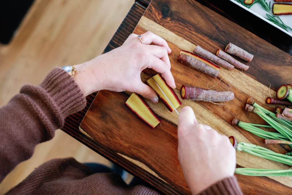 overhead shot of hands cutting brightly colored carrots on wooden cutting board.