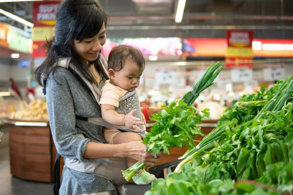 woman holding a baby in a carrier on her chest as she shops for produce