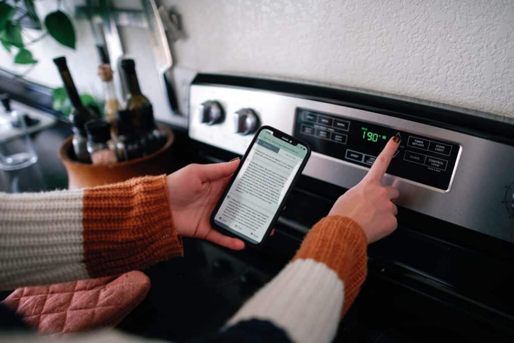 over the shoulder shot of a woman holding a phone with recipe directions and updating the temp on an oven