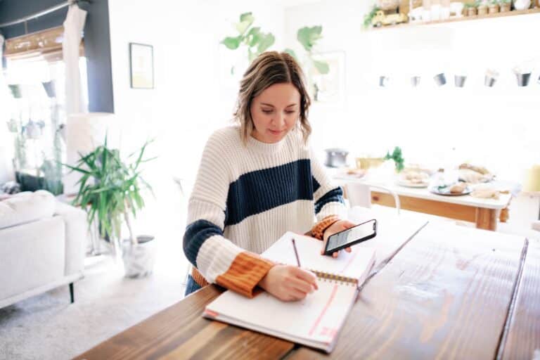 woman sitting at a dining room table writing on a planner with a cell phone in her hand