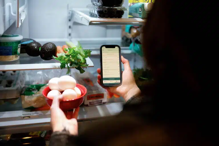 over the shoulder image of a person looking in a fridge with a phone in hand and eggs placing eggs in the fridge