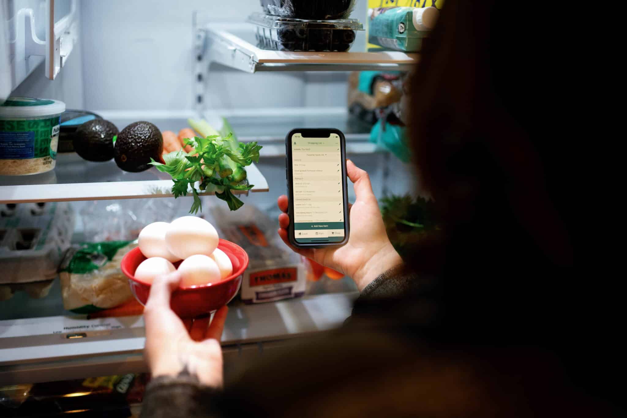 over the shoulder image of a person looking in a fridge with a phone in hand and eggs placing eggs in the fridge