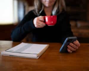 cropped image of woman drinking from a red coffee mug, holding her phone with a planner next to her