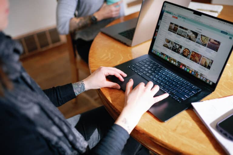 over the shoulder view of woman viewing recipes on her laptop at a wood table