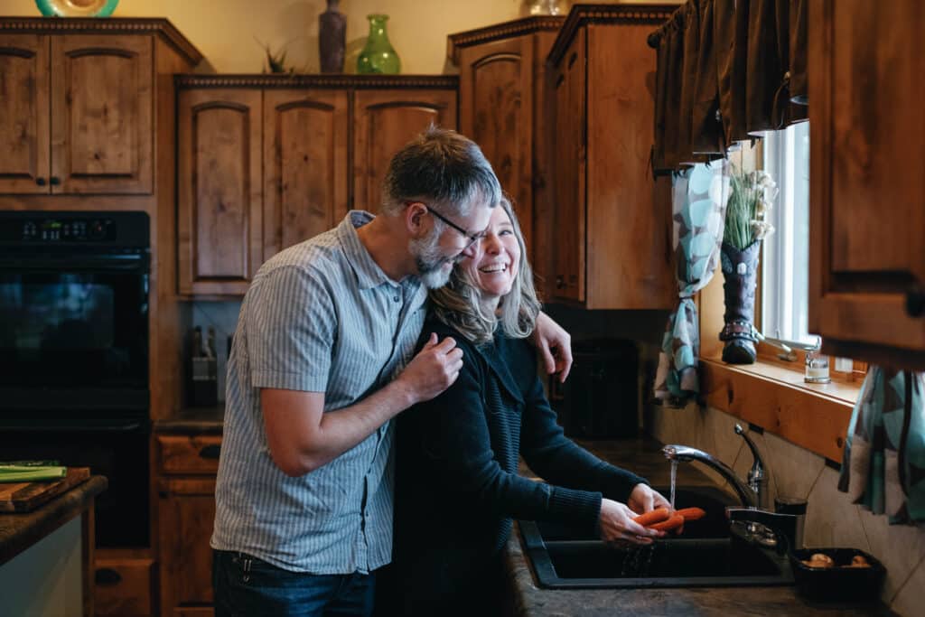 Clint hugging Lisa as she cleans carrots in a kitchen sink