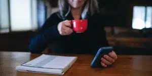 cropped image of a woman drinking from a red mug with a phone in her hand.