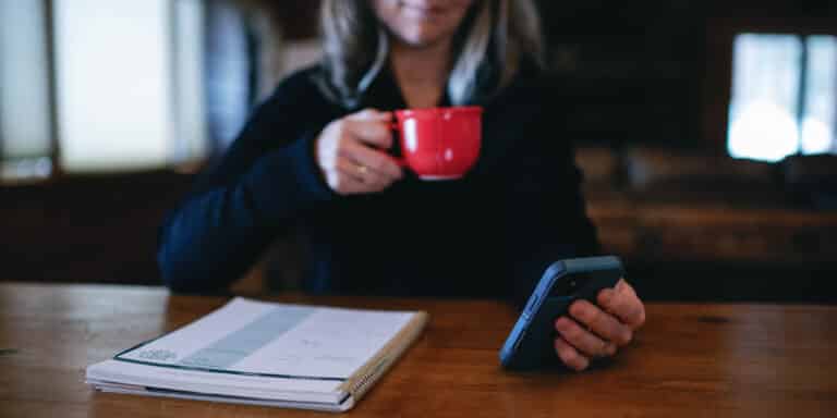 cropped image of a woman drinking from a red mug with a phone in her hand.